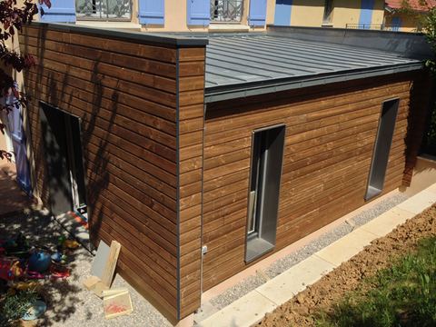 Extension cubique en bardage bois foncé et toiture zinc, accolée à une maison aux volets bleus